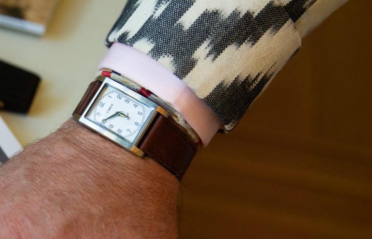 Ceas dreptunghiular WM Brown x Timex cu cadran alb și curea maro din piele purtat pe încheietura mâinii, lângă manșetă roz și sacou cu model alb-negru.