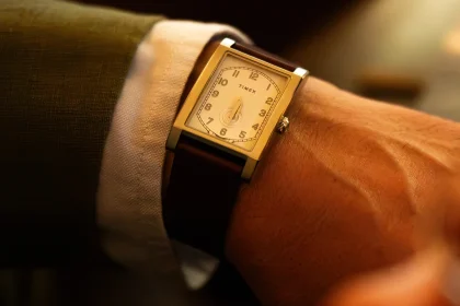 Prim-plan cu ceas WM Brown x Timex cu carcasă rectangulară și curea maro din piele, purtat pe încheietura mâinii lângă manșeta unei cămăși, iluminat cald.