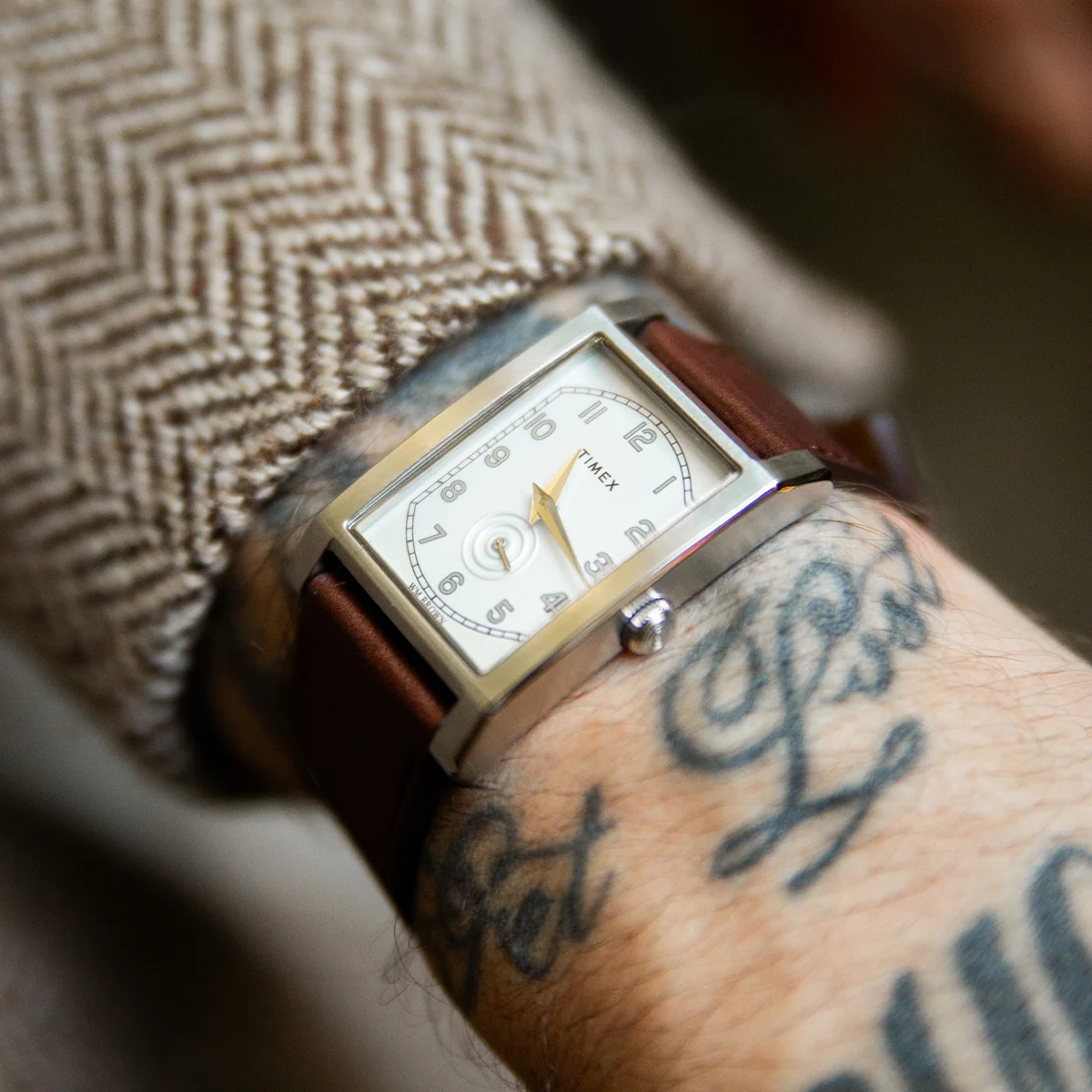 14341 TX TC 26 FGI desktop TWG069000 2 Ceas Timex WM Brown x Timex cu cadran alb, carcasă rectangulară argintie și curea maro din piele, purtat pe un încheietura cu tatuaj.