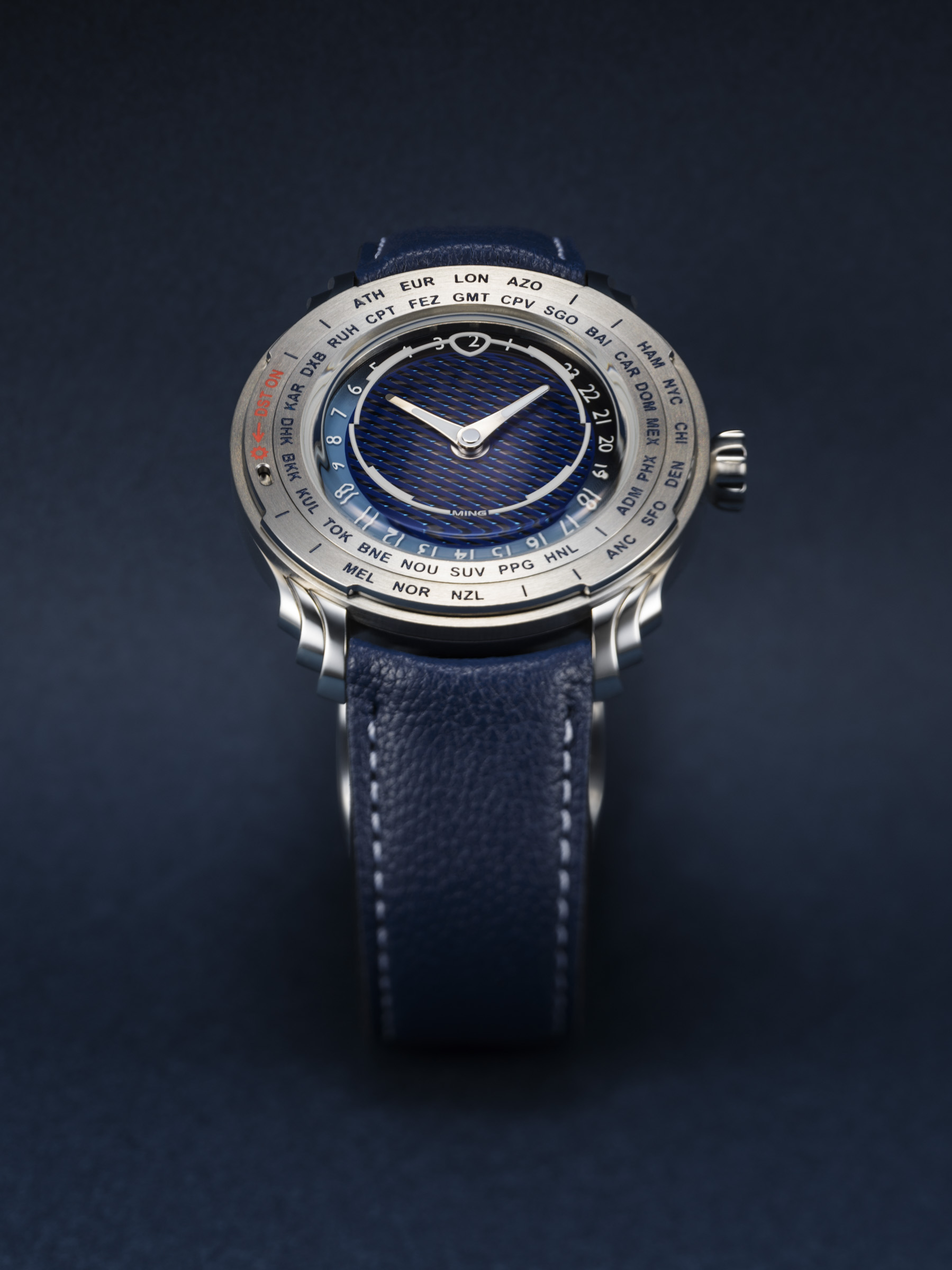 6a7ce4f4345a5c0ee008629b 907X B0008963 Ceas MING 57.05 Comet cu cadran albastru texturat, carcasă din metal argintiu, bezel cu nume de orașe, curea albastră din piele cu cusături albe.