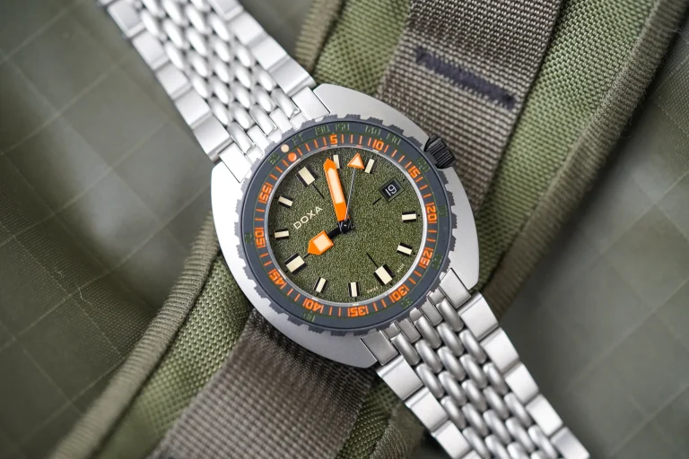 Ceas Doxa Sub 300 Beta cu cadran verde texturat, indicatoare oranj și brățară metalică, așezat pe material textil kaki.