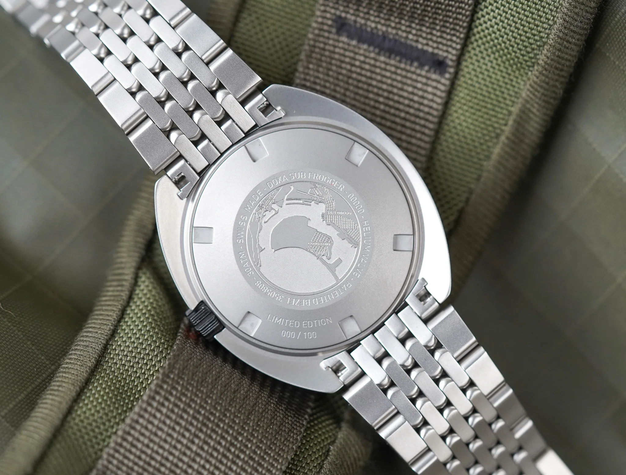 DSC02907 21a35fd8 46a3 4990 aadf c401e1b944d8 Spatele carcasei ceasului Belmont Watches X Doxa Sub 300 Beta Frogger cu gravură și brățară metalică, pe fundal textil verde kaki și maro.
