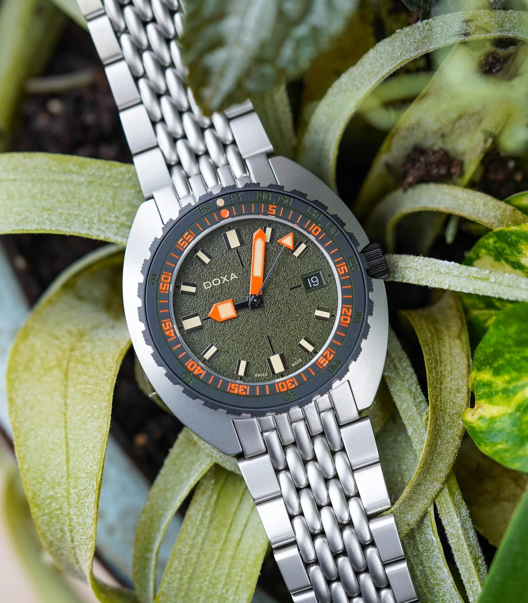 DSC02911 Ceas Doxa Sub 300 Beta cu cadran verde, indicatori și ace oranj, curea metalică, poziționat pe plante verzi.