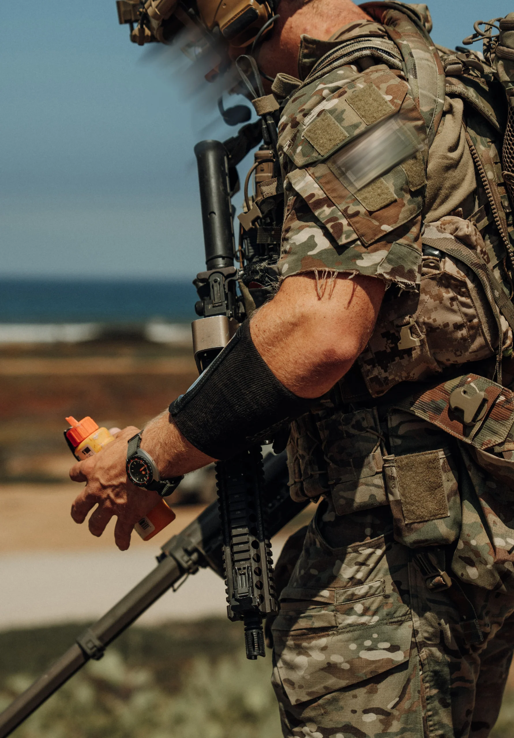 Frogger Navy Seal 7 scaled Soldat în echipament camuflat ținând o armă și o sticlă portocalie, cu ceas negru la încheietura mâinii stângi, pe fundal de coastă și mare.