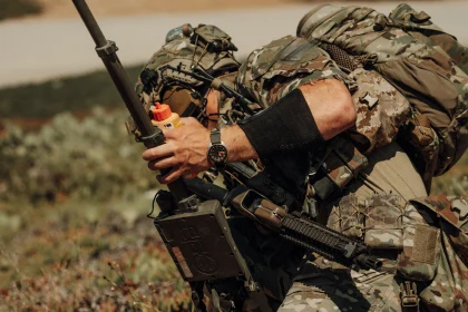 Soldat în echipament de camuflaj, cu ceasul Belmont Watches X Doxa Sub 300 Beta 'Frogger' la mână, ținând un dispozitiv și un container portocaliu în peisaj deschis.