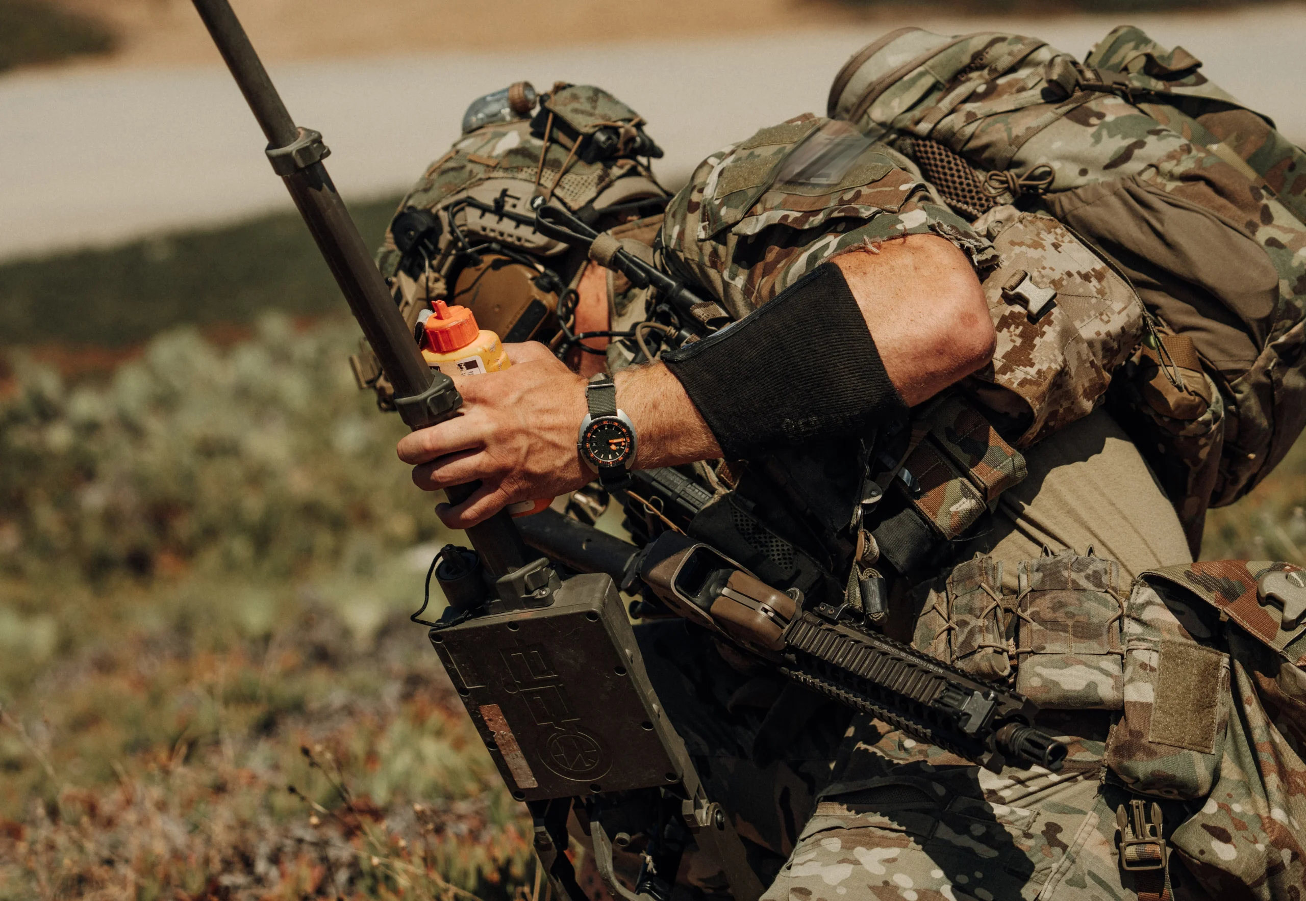 Frogger Navy Seals Image scaled Soldat în echipament de camuflaj, cu ceasul Belmont Watches X Doxa Sub 300 Beta 'Frogger' la mână, ținând un dispozitiv și un container portocaliu în peisaj deschis.