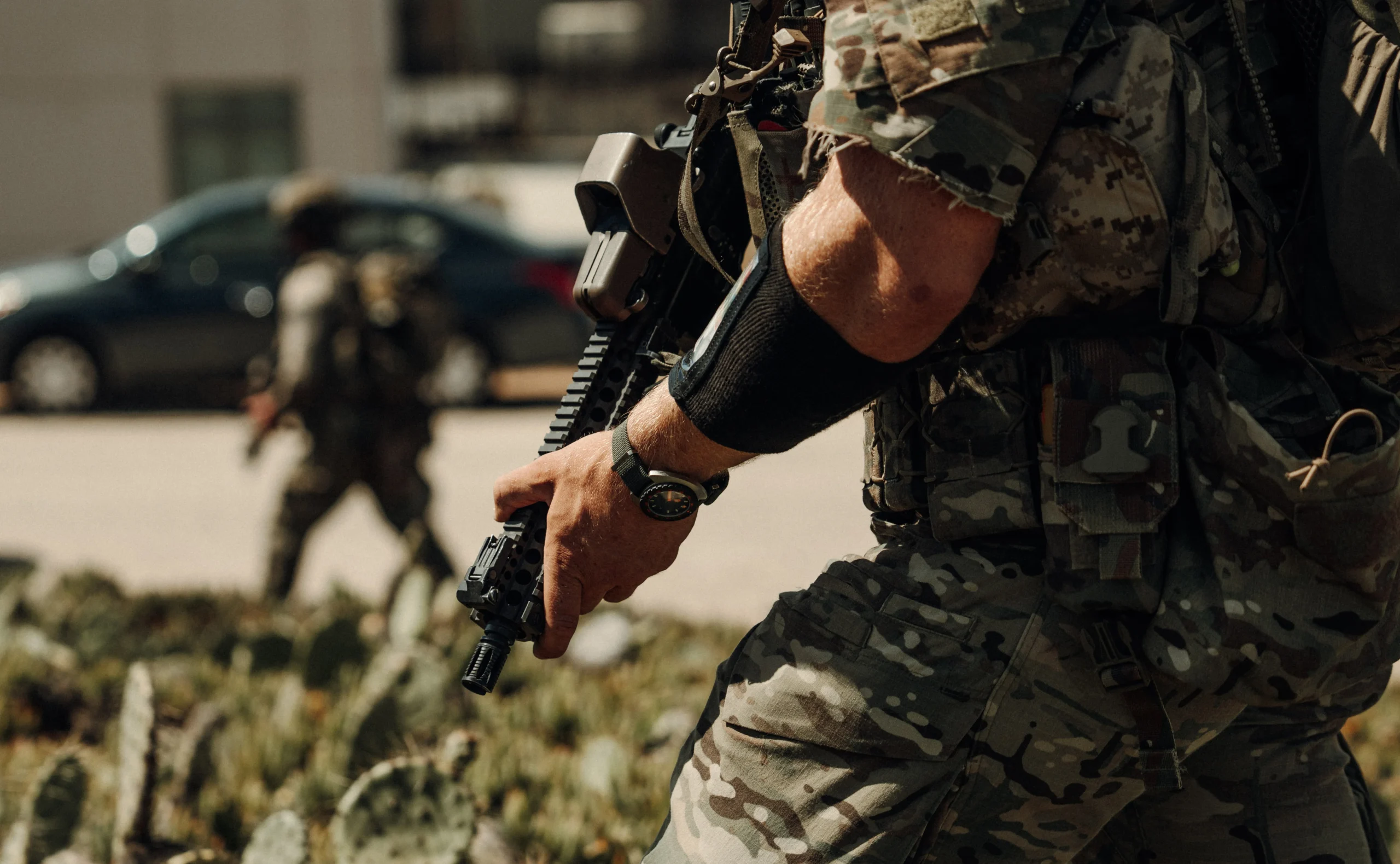 Navy Seal Frogger 3 2ce86ba4 e5c1 41c1 9a32 eb9b9006f50d scaled Soldat în uniformă camuflată care ține o armă și poartă un ceas pe încheietura mâinii, cu un alt soldat blurat în fundal.