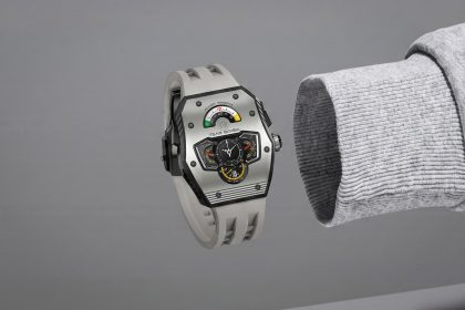 Ceas cu carcasă argintie și curea gri deschis, afișaj multifuncțional cu indicator de rezervă de putere și cadrane suplimentare colorate, purtat pe încheietură cu mânecă gri deschisă.