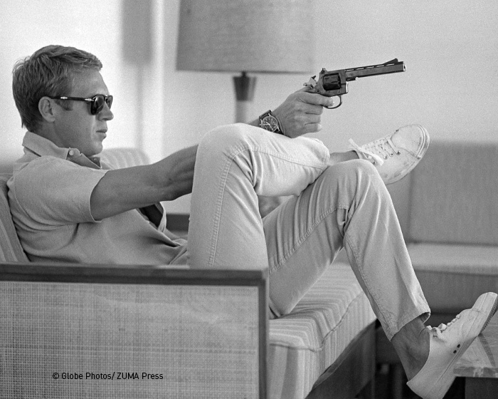 hanhart Steve McQueen website 1 700x560px 1 Bărbat purtând ochelari de soare, stând pe canapea cu picioarele ridicate, ținând un pistol în mână și având un ceas la mână.
