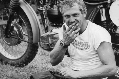 Portret alb-negru al unui bărbat tânăr cu tricou Triumph, șezând lângă o motocicletă clasică, purtând un ceas pe încheietura mâinii.
