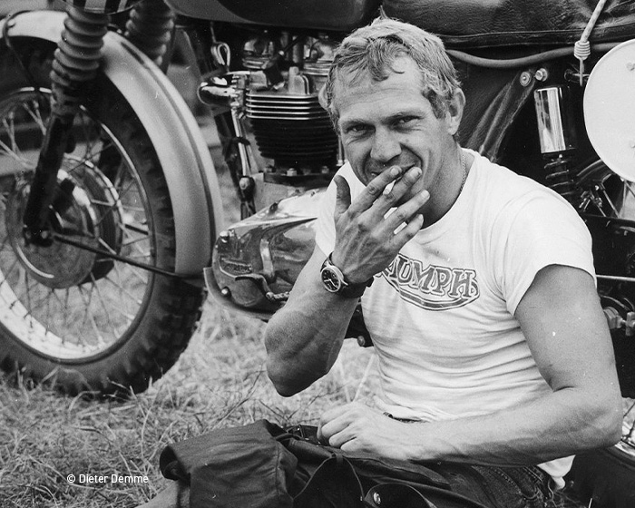 hanhart Steve McQueen website 3 700x560px 2 Portret alb-negru al unui bărbat tânăr cu tricou Triumph, șezând lângă o motocicletă clasică, purtând un ceas pe încheietura mâinii.