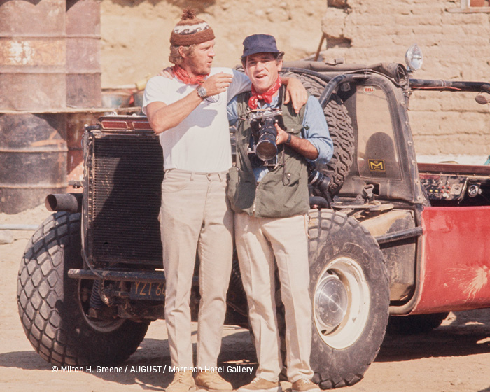 hanhart Steve McQueen website 6 700x560px 1 Două bărbați îmbrăcați casual stau îmbrățișați lângă un vehicul off-road vechi, unul poartă o pălărie de lână, celălalt o șapcă și are un aparat foto la gât.