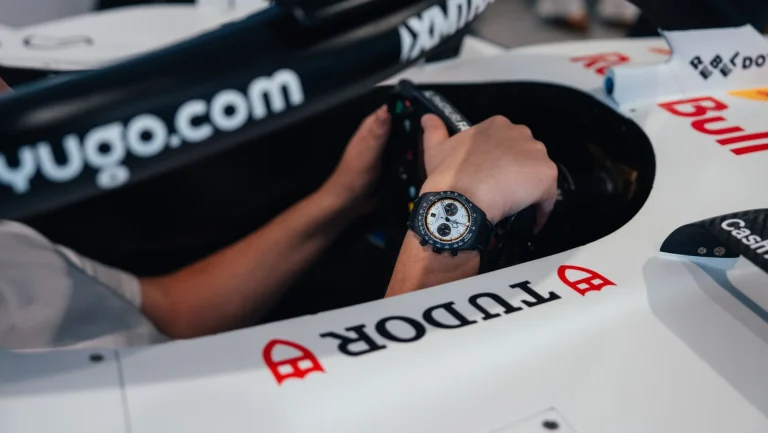 Mana unui pilot Formula 1 cu ceas Tudor negru cu cadran alb si subcadrane, tinand volanul unei masini de curse albe cu logo Tudor si Red Bull.