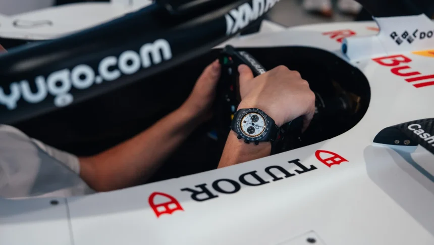 Mana unui pilot Formula 1 cu ceas Tudor negru cu cadran alb si subcadrane, tinand volanul unei masini de curse albe cu logo Tudor si Red Bull.
