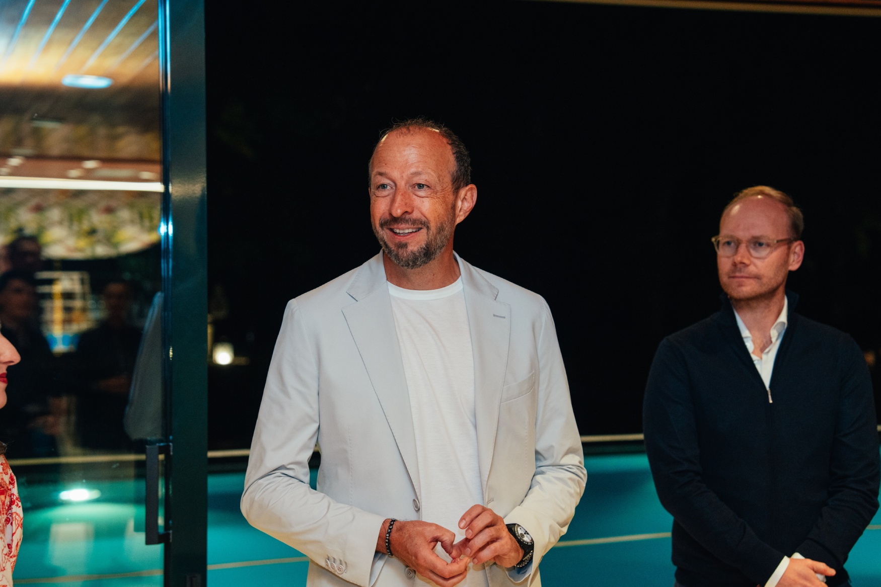 tudor vcarb f1 tour 4 Bărbat cu sacou alb și tricou alb, zâmbind într-un cadru de seară lângă o piscină, alături de alt bărbat cu jachetă albastră închisă.